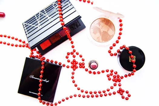 Moscow, 2021. White powder and bronzer, packages with eye shadow, lipstick, blush and a brush of different brands lie on a white background wrapped in red beads. High quality photo
