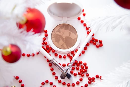 A white pillow with powder and bronzer lies on a white background among a white fluffy Christmas tree with red toys, thin makeup brushes and bright red beads lie in front. High quality photo