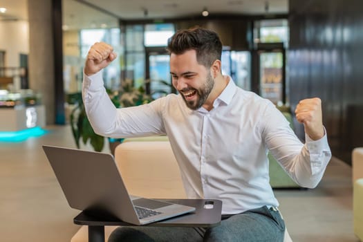 Caucasian bearded businessman working on laptop shocked by sudden victory, game winning lottery goal achievement, good career news. Excited guy freelancer clenching fists sitting in modern hotel lobby