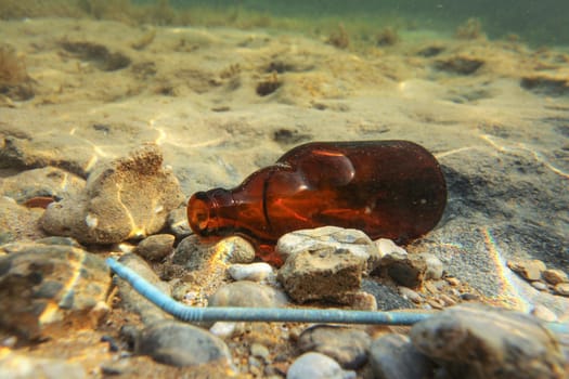 Small brown beer bottle and blue plastic straw on sandy sea bottom. Underwater photo, ocean littering concept.