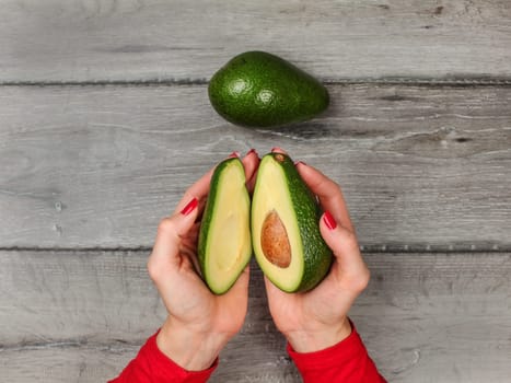 Tabletop view, young woman hands holding two halves of avocado cut in half, over gray wood desk, whole green pear next to it.