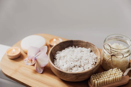Preparation for hotel spa treatment or home bath procedure. White washbasin in bathroom with accessories on tray. Burning candles, washcloth, soap, foot brush, bottle with sea salt, orchid flower