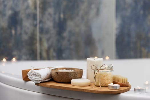 Preparation for hotel spa treatment, home bath procedure. White washbasin in bathroom, accessories on tray. Burning candles, soap, foot brush, towel, glass bottle with sea salt, orchid flower