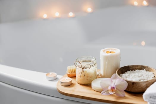 Preparation for hotel spa treatment or home bath procedure. White washbasin in bathroom with accessories on tray. Burning candles, washcloth, soap, foot brush, bottle with sea salt, orchid flower