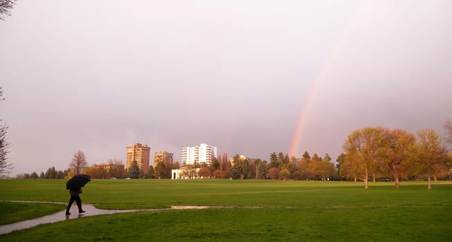 The sun peaks through for a moment lighting the way and producing a rainbow near a person walking with umbrella