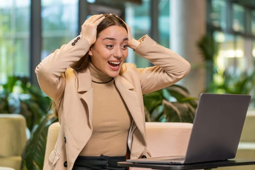 Happy businesswoman girl using laptop computer typing browsing, celebrating success victory, winning lottery casino jackpot achievement, play game, good positive news, triumph in modern office lobby