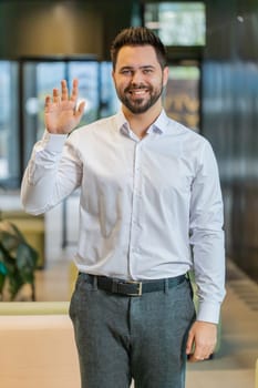 Hello. Happy young Caucasian businessman employee waves hand hi gesture greeting welcomes someone, online webinar, remote distance conversation. Entrepreneur man in shirt standing in office lobby.