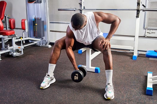 An African American man shakes his biceps while sitting on a bench in the gym. High quality photo