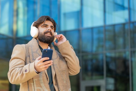 Happy Arabian Indian businessman putting on wireless headphones choosing, listening favorite energetic music in smartphone outdoors. Smiling man guy walking passes by urban city street. Lifestyles.