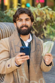 Happy rich Indian businessman tourist using smartphone and counting money dollar cash banknotes sitting on bench on downtown city street. Success business career, lottery winner, big income, wealth
