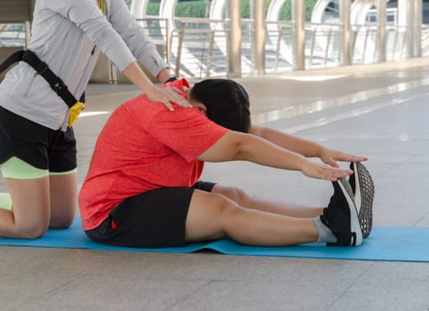 Instructor supporting overweight woman while doing stretching exercises outdoors. Plus size young woman sitting and touching her toes on yoga mat