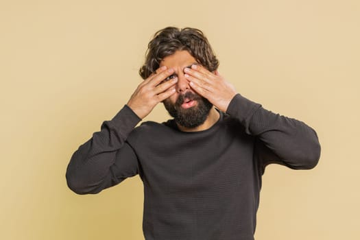 Dont want to look at this, awful. Indian shocked bearded man closing eyes with hands showing stop gesture, confused shy scared to watch. Young adult guy boy isolated alone on beige studio background.