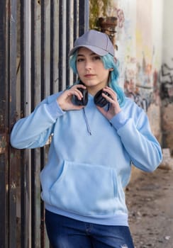 Teenager wearing light blue oversize hoodie and baseball cap looking into camera. Blue haired teen girl with wireless headphones stays outdoor near a gate. Clothing and baseball cap mockup
