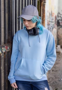 Teenager wearing light blue oversize hoodie and baseball cap looking down. Blue haired teen girl with wireless headphones stays outdoor near a gate. Clothing and baseball cap mockup