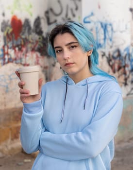 Teenage girl in light blue oversize hoodie with coffee to go and looking into the cameraBlue haired teen girl staying outdoors near graffiti wall. Clothing mockup. Hipster and adolescence concept