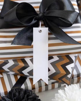 Present with blank vertical gift tag and black bow close up. Gift boxes wrapped in Striped and chevron geometric paper near black and white decor. Christmas, New Year, Birthday, Anniversary label mockup