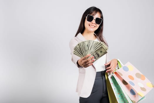 Woman with glasses confident shopper smile holding online shopping bags multicolor and dollar money banknotes on hand, excited happy Asian young female person studio shot isolated on white background