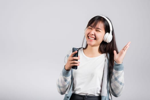 Woman excited smiling listening to music radio in bluetooth headphones and singing song on smartphone studio shot isolated white background, happy Asian young female Karaoke online app in mobile phone