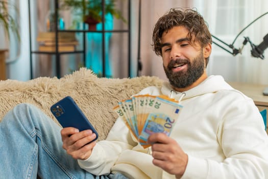 Planning budget. Rich happy Indian man using smartphone calculator app count euro cash, calculate domestic bills at home. Guy satisfied of income earnings win saves money for planned vacation gifts.