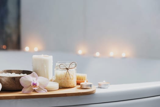 Preparation for hotel spa treatment or home bath procedure. White washbasin in bathroom with accessories on tray. Burning candles, washcloth, soap, foot brush, bottle with sea salt, orchid flower