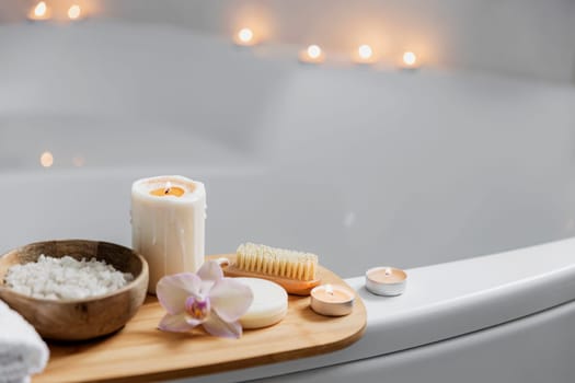 Preparation for hotel spa treatment, home bath procedure. White washbasin in bathroom, accessories on tray. Burning candles, soap, foot brush, towel, glass bottle with sea salt, orchid flower