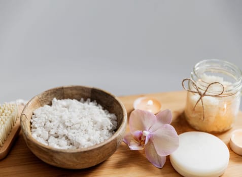 Preparation for hotel spa treatment or home bath procedure. White washbasin in bathroom with accessories on tray. Burning candles, washcloth, soap, foot brush, bottle with sea salt, orchid flower