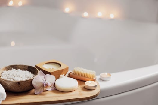 Preparation for hotel spa treatment or home bath procedure. White washbasin in bathroom with accessories on tray. Burning candles, washcloth, soap, foot brush, bottle with sea salt, orchid flower