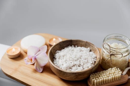 Preparation for hotel spa treatment or home bath procedure. White washbasin in bathroom with accessories on tray. Burning candles, soap, foot brush, glass bottle with sea salt, orchid flower
