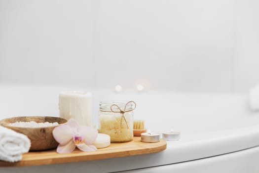 Preparation for hotel spa treatment, home bath procedure. White washbasin in bathroom, accessories on tray. Burning candles, soap, foot brush, towel, glass bottle with sea salt, orchid flower