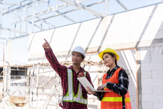Asian engineer foreman worker man and woman working at building construction site use laptop and talking with radio, engineering hold computer and radio discuss and control worker employee to building