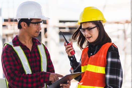 Two Asian engineer foreman architect worker man and woman talking at building construction site, engineering hold radio discussion operate project and control worker employee to building construction