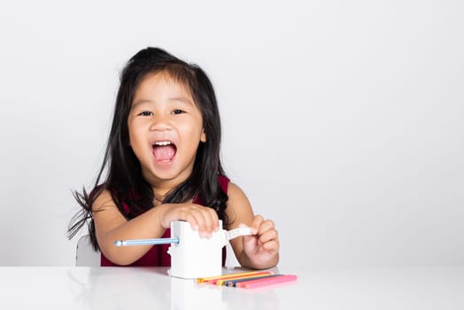 Little cute kid girl 3-4 years old smile using pencil sharpener while doing homework in studio shot isolated on white background, Asian children preschool sharpening color pencils, education concept