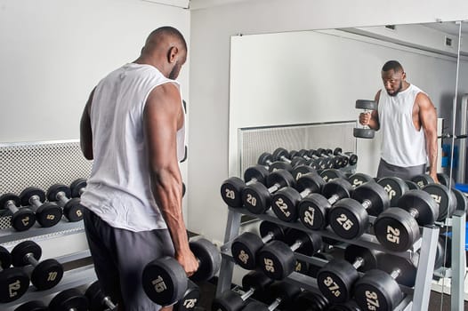 An African American man shakes his biceps with dumbbells while standing in front of a mirror. High quality photo