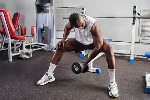 An African American man shakes his biceps while sitting on a bench in the gym. High quality photo