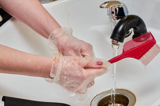 Women's hands in hair coloring gloves wash a comb under a stream of water in a white sink. High quality photo