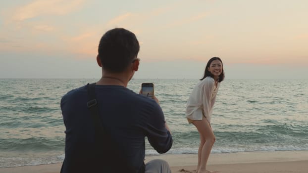 Happy man using smartphone camera making photo to woman at the beach and having fun together on beach in holiday vacation time at sunset, Young Asian woman and man in summer travel outdoor