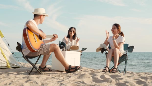 Happy friend have fun playing guitar and clap in camp they smiling together in holiday on sand beach near camping tent vacation time at sunset, Young Asian group woman and man in summer travel outdoor