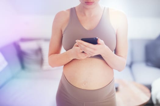 Closeup photo of pregnant female belly. Woman holding and using mobile smart phone app at home interiors. Pregnancy, technology, online shopping, preparation and expectation concept.