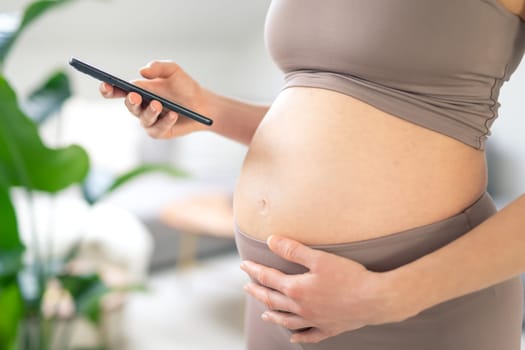 Closeup photo of pregnant female belly. Woman holding and using mobile smart phone app at home interiors. Pregnancy, technology, online shopping, preparation and expectation concept.