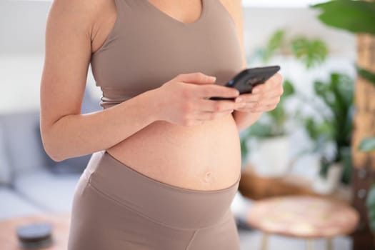 Closeup photo of pregnant female belly. Woman holding and using mobile smart phone app at home interiors. Pregnancy, technology, online shopping, preparation and expectation concept.