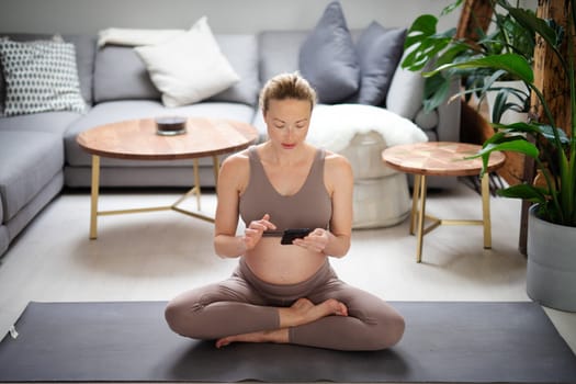 Pregnancy and technoloy supported healthy lifestyle concept. Cheerful happy pregnant woman using smart phone application while exercising on yoga mat on living room floor at home