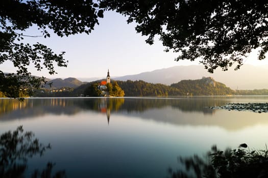 Lake Bled with a church on an island at sunrise through window of branches. Peaceful scenery with a religion building in the middle of nature illuminated with morning light with copy space.