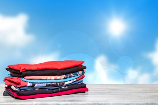 Stack colorful clothes. Closeup of a pile of colorful t-shirts or shirts on a bright table against abstract blurred sunny blue sky background. Space for your product display montage. Summer fashion.