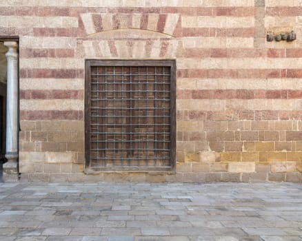 Daylight shot of wooden window with wrought iron grid in a wall decorated with beige and red stones