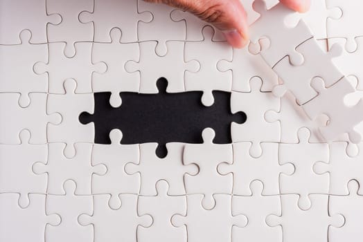 Top view flat lay of hand-holding last piece white paper jigsaw puzzle game last pieces put to place for solve problem complete mission, studio shot on a black background, quiz calculation concept