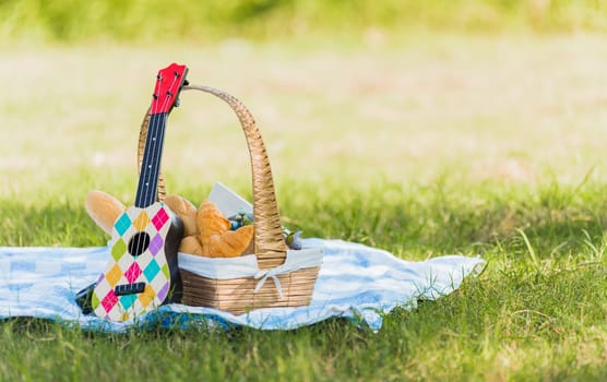Picnic wattled basket with bread food and fruit, Ukulele, a retro camera on blue cloth in green grass garden with copy space at sunny summertime