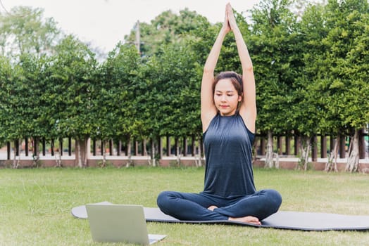 Asian young woman using laptop during online video training practicing yoga outdoors in meditate pose sitting on green grass in nature a field garden park, Stretching health care concept