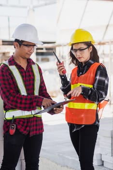 Two Asian engineer foreman architect worker man and woman talking at building construction site, engineering hold radio discussion operate project and control worker employee to building construction