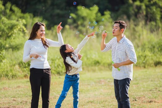 Happy Asian lifestyle family mother, father and little cute girl child having fun together and enjoying outdoor play blowing soap bubbles in the garden park on a sunny day, summertime