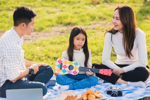 Happy Asian young family father, mother and children having fun and enjoying outdoor together sitting on the grass party with playing Ukulele during a picnic in the garden park on a sunny day